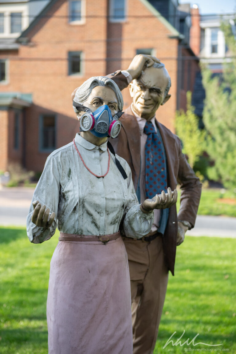 A photograph of statues of a man and woman; the woman wears a surgical mask and then man looks at her