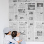 A photograph of two children sitting on the floor and gazing at the multiple newspapers covering a white wall. The newspapers are pertaining to the year 2020 and the news regarding COVID-19.