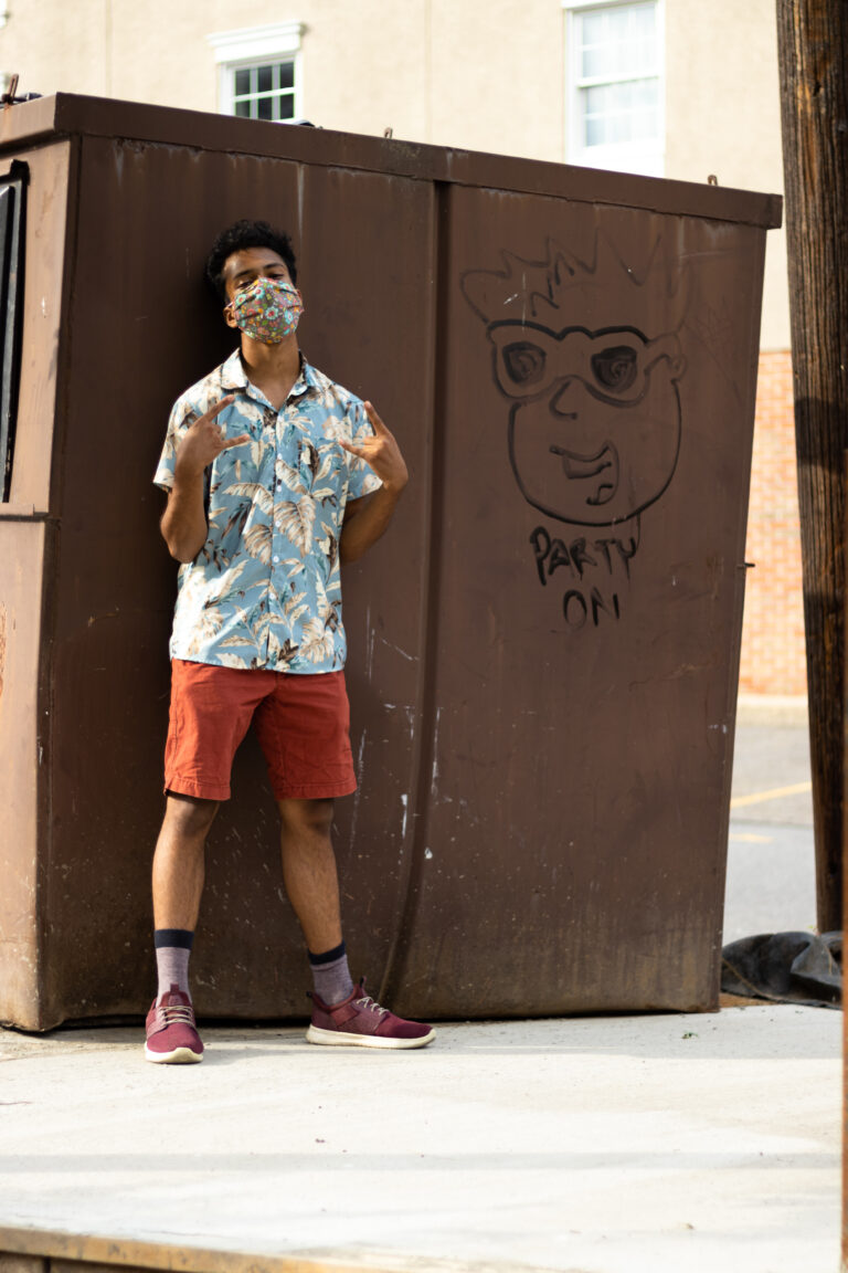Man with a mask on standing in front of a graffitied dumpster