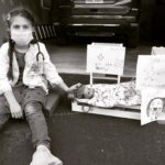 A black and white photograph of a girl sitting next to a toy driveway clinic with a doll in a bed and signs stating